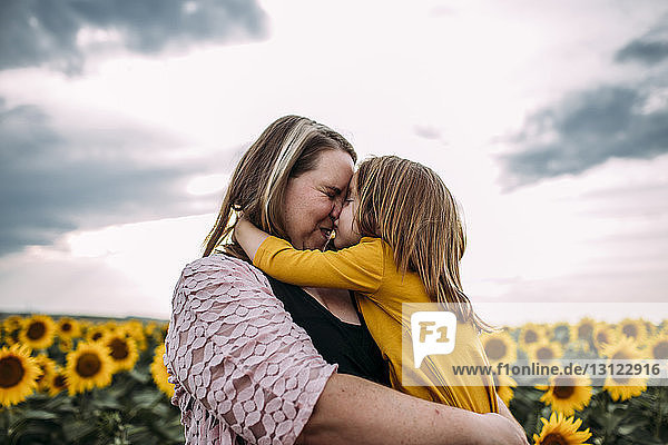 Mutter und Tochter umarmen sich  während sie in einer Sonnenblumenfarm gegen den Himmel stehen