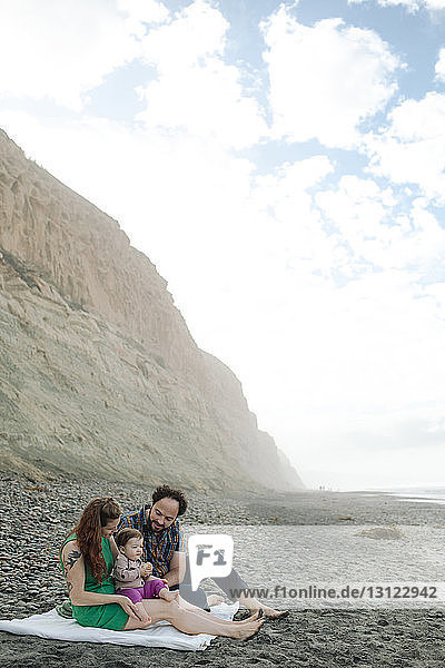 Family relaxing at beach by mountain against cloudy sky