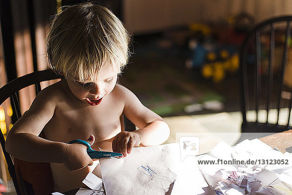 High angle view of shirtless boy cutting papers with scissors while sitting on chair at home