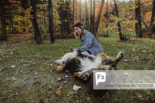 Happy woman playing with dog lying on grassy field in forest during autumn