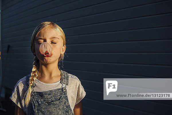 Girl with eyes closed blowing bubble gum while standing by wall