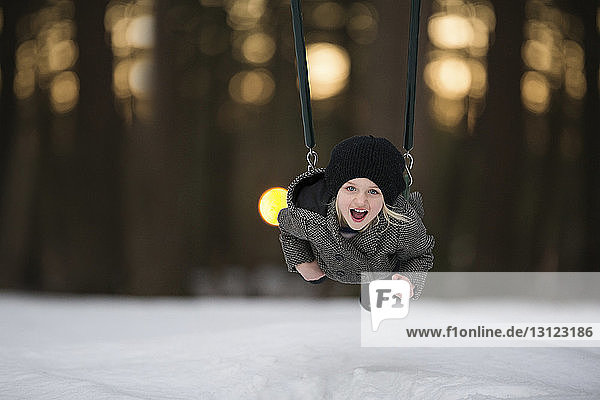 Portrait of cheerful girl playing on swing at snow covered park