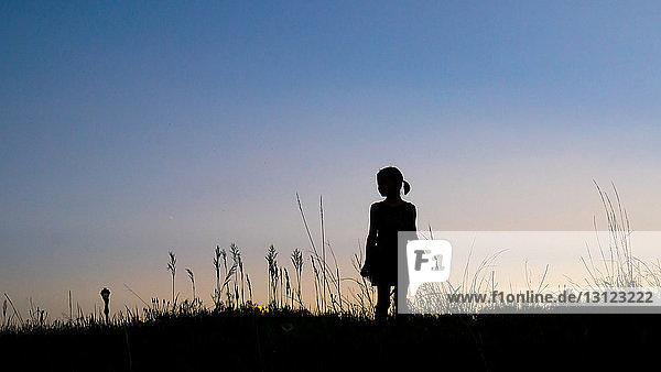 Silhouettenmädchen steht in der Dämmerung auf Grasfeld vor klarem Himmel