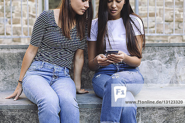 Female friends using mobile phone while sitting on steps against wall