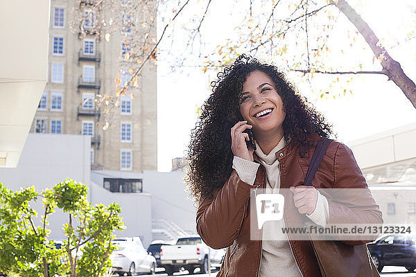 Niedrigwinkelansicht einer glücklichen Frau  die mit einem Smartphone telefoniert  während sie in der Stadt gegen den Himmel steht