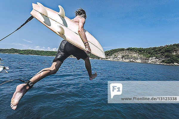 Mann mit Surfbrett springt am sonnigen Tag gegen den Himmel ins Meer