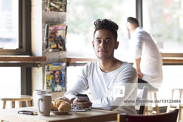 Portrait of young man holding mobile phone having croissant and coffee at cafe