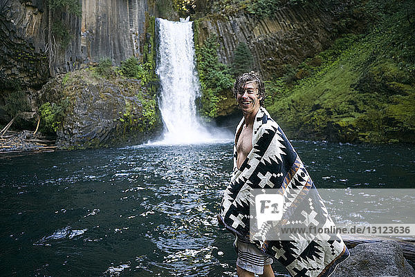 Porträt eines glücklichen Mannes im Tuch  der am Wasserfall im Wald steht
