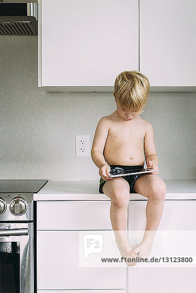 Junge ohne Hemd benutzt Tablet-Computer  während er zu Hause auf dem Küchentisch sitzt