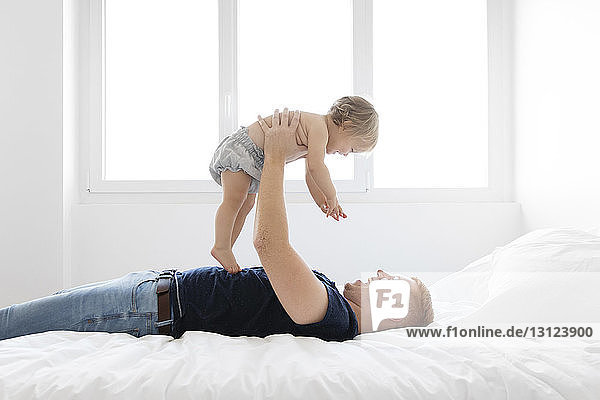 Side view of happy father lifting shirtless daughter while lying on bed at home