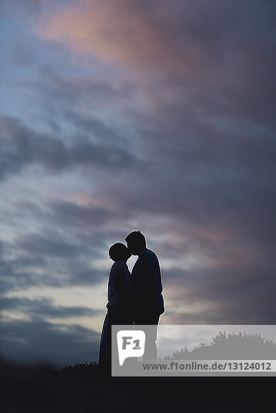 Silhouettenpaar küsst sich  während es bei Sonnenuntergang auf dem Feld vor bewölktem Himmel steht