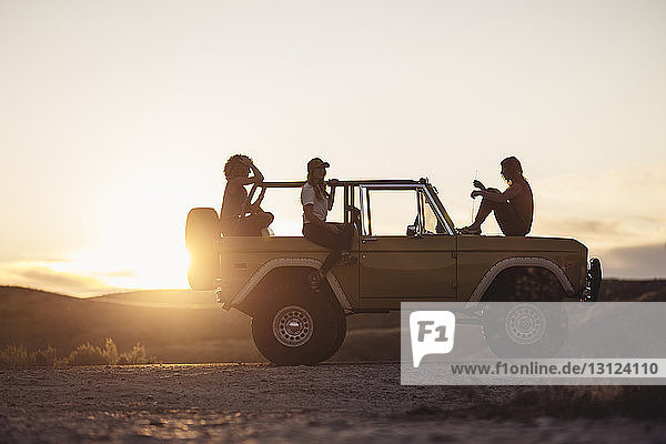 Freundinnen sitzen bei Sonnenuntergang auf Geländewagen gegen den Himmel