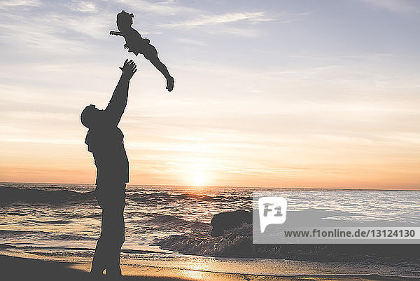 Seitenansicht des Silhouettenvaters  der seine Tochter wirft  während er bei Sonnenuntergang am Strand gegen den Himmel steht
