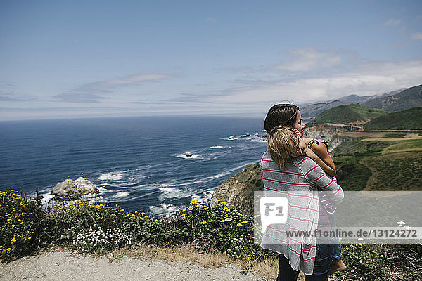 Mother carrying daughter while standing on cliff against sea