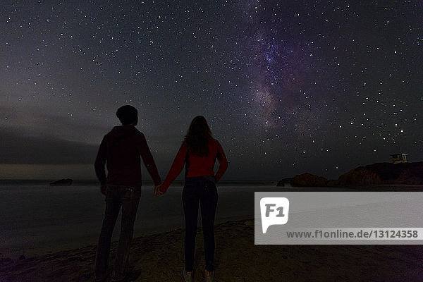 Rückansicht eines Paares  das sich an den Händen hält  während es den nächtlichen Sternenhimmel betrachtet