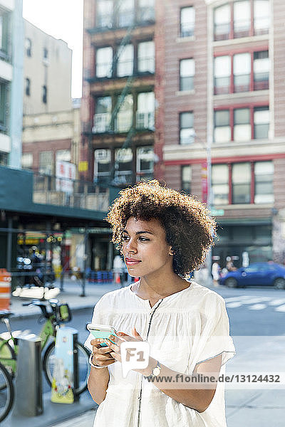 Woman looking away while holding smart phone in city