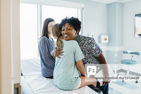 Kinderarzt umarmt Mädchen  das von der Mutter auf dem Untersuchungstisch im Krankenhaus sitzt
