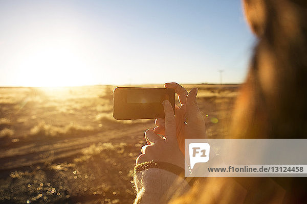 Frau fotografiert Feld mit Smartphone bei Sonnenuntergang