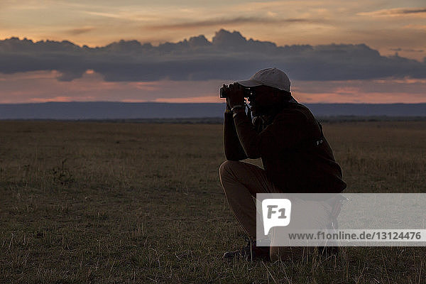 Mann in voller Länge mit Fernglas  während er bei Sonnenuntergang auf einem Feld im Serengeti-Nationalpark kniet