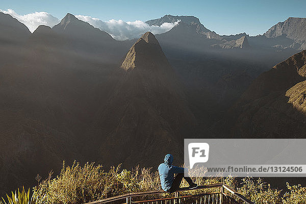 Rückansicht eines Mannes  der Berge betrachtet  während er auf einem Beobachtungspunkt sitzt