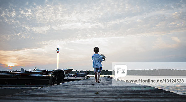 Rückansicht eines Jungen  der bei Sonnenuntergang auf dem Pier vor bewölktem Himmel läuft