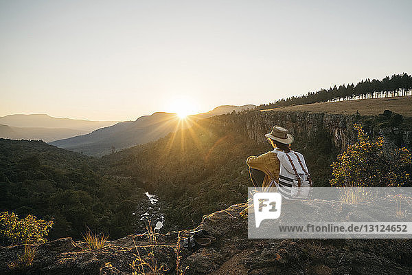 Side view of woman with backpack looking at view while sitting on mountain against sky during sunset