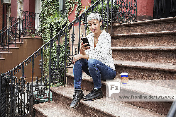 Frau benutzt Mobiltelefon  während sie mit Kaffeetasse auf der Treppe sitzt