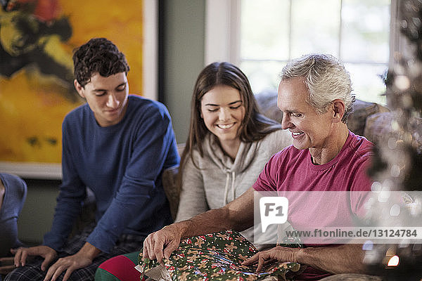 Grossvater packt Weihnachtsgeschenk aus  während er mit den Enkelkindern zu Hause sitzt