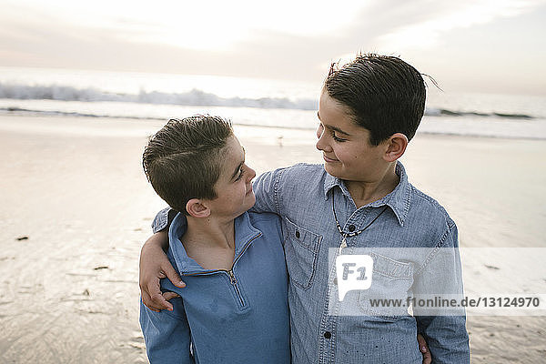Smiling brothers standing at beach against sky during sunset