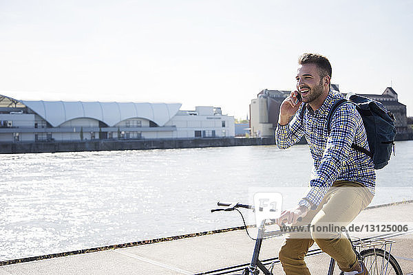 Happy man talking on phone while cycling by canal against clear sky