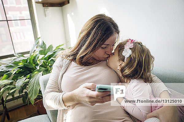 Mutter küsst Tochter  während sie zu Hause ein Smartphone in der Hand hält