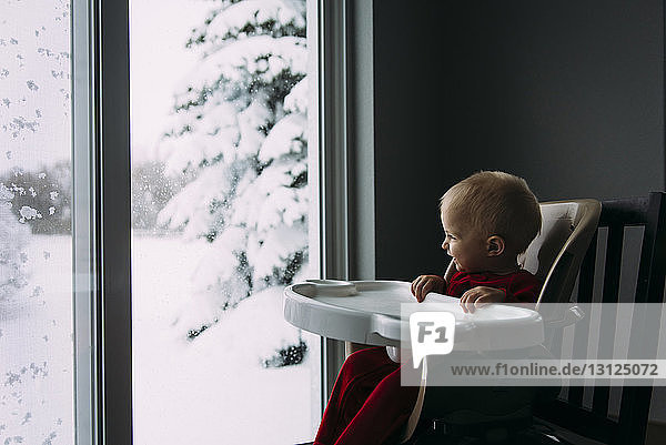 Glücklicher kleiner Junge  der im Winter zu Hause auf einem Hochstuhl sitzend durch das Fenster schaut