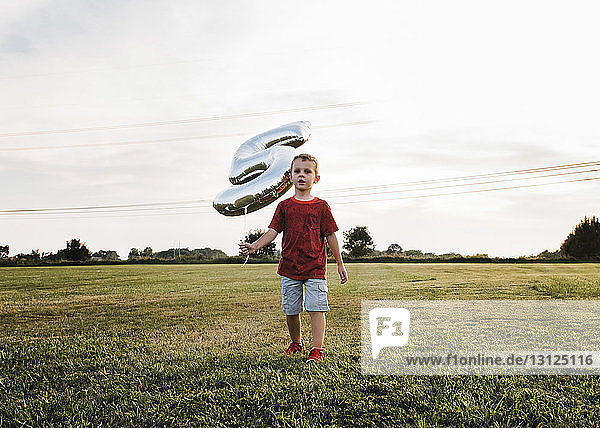 Junge hält Ballon Nummer 5 in der Hand  während er auf Grasfeld gegen den Himmel läuft