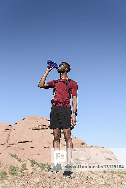 Niedrigwinkelaufnahme eines männlichen Wanderers  der Wasser trinkt  während er auf einer Felsformation vor klarem blauen Himmel steht