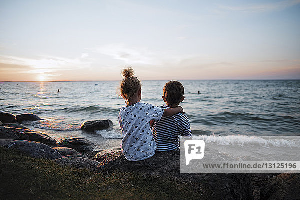 Rear view of siblings sitting on rocks by Lake Simcoe against sky during sunset