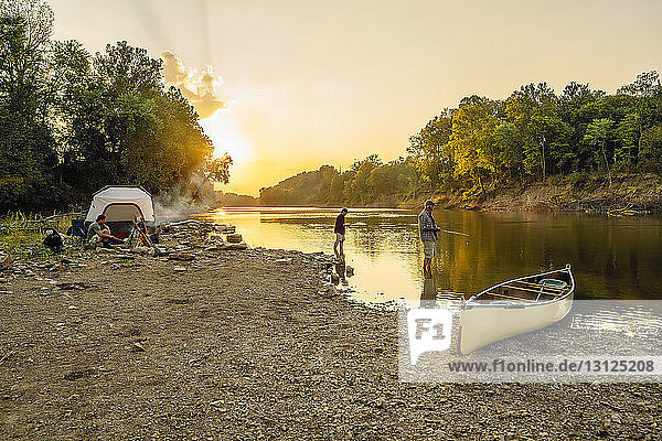 Junger Mann sitzt auf dem Campingplatz  während Freunde bei Sonnenuntergang im See angeln