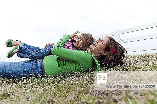 Cheerful mother embracing daughter while lying on grass against clear sky