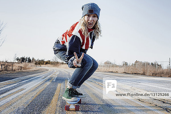 Portrait of playful woman gesturing peace sign while skateboarding on road during winter