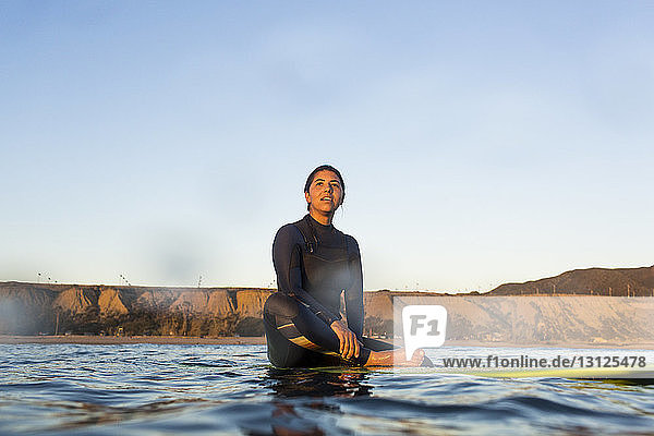 Junge Frau sitzt auf einem Surfbrett im Meer vor klarem Himmel