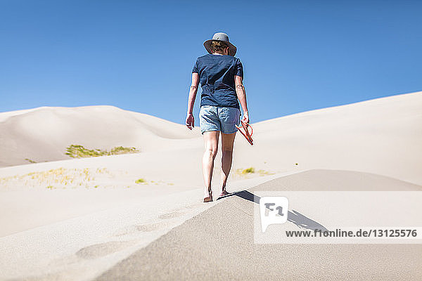 Rear view of woman holding flip-flop while walking on sand at Great Sand Dunes National Park during sunny day