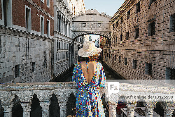 Rückansicht einer Frau mit Hut  die an einem Geländer auf einer Brücke inmitten von Gebäuden in der Stadt steht