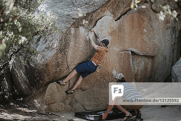 Mann hält Sicherheitsmatte  während ein Freund im Wald am Fels klettert