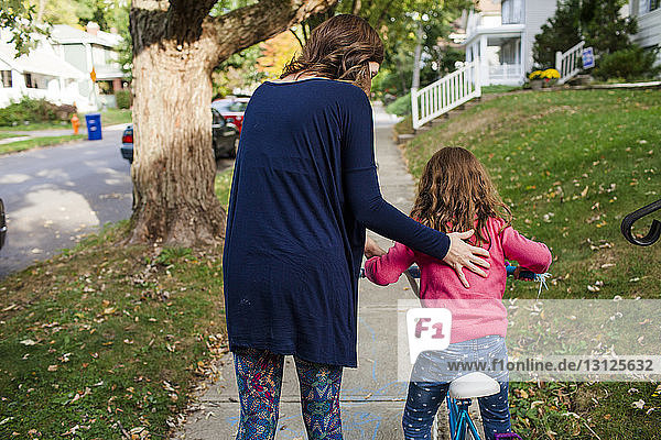 Rückansicht der Mutter  die der Tochter im Herbst beim Fahrradfahren auf dem Fußweg hilft