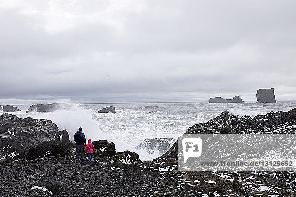 Rear view of father with daughter looking at sea while standing on rock by Dyrholaey Iceland against cloudy sky