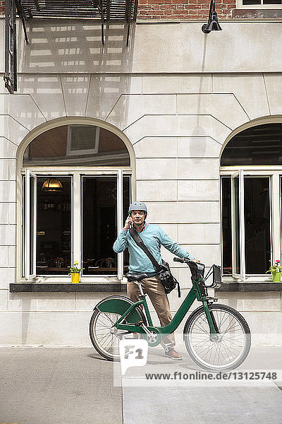 Reifer Mann benutzt Smartphone  während er mit Fahrrad gegen Gebäude steht
