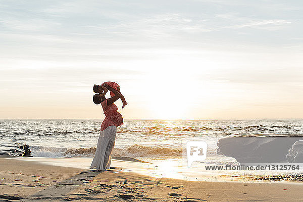 Seitenansicht einer schwangeren Mutter  die ihre Tochter hochhebt  während sie bei Sonnenuntergang am Strand gegen den Himmel steht