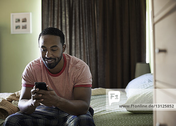 Smiling man using phone while sitting on bed at home