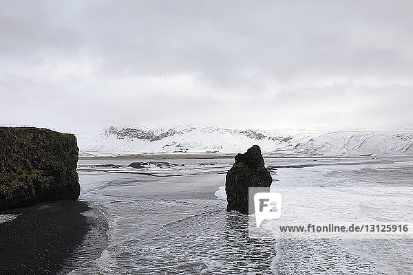 Landschaftliche Ansicht von Dyrholaey Island und Arnardrangur Felsen gegen bewölkten Himmel im Winter