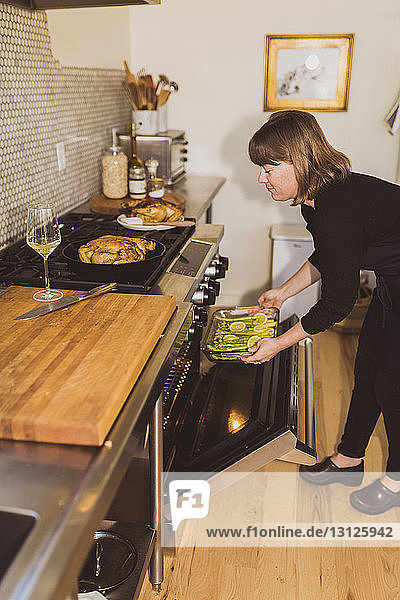 Woman keeping asparagus with lemons on tray in oven