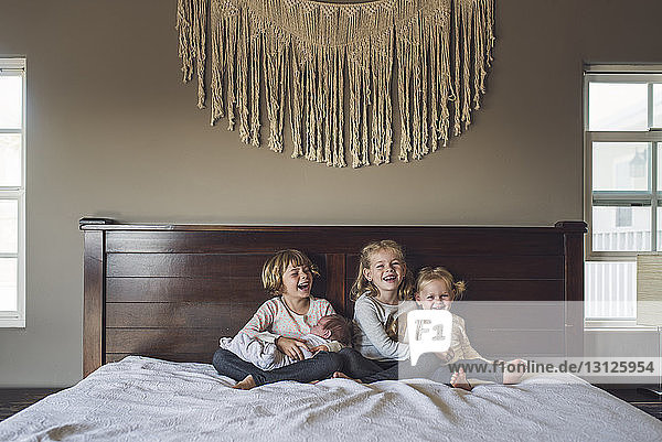 Cheerful sisters with baby girl sitting on bed at home